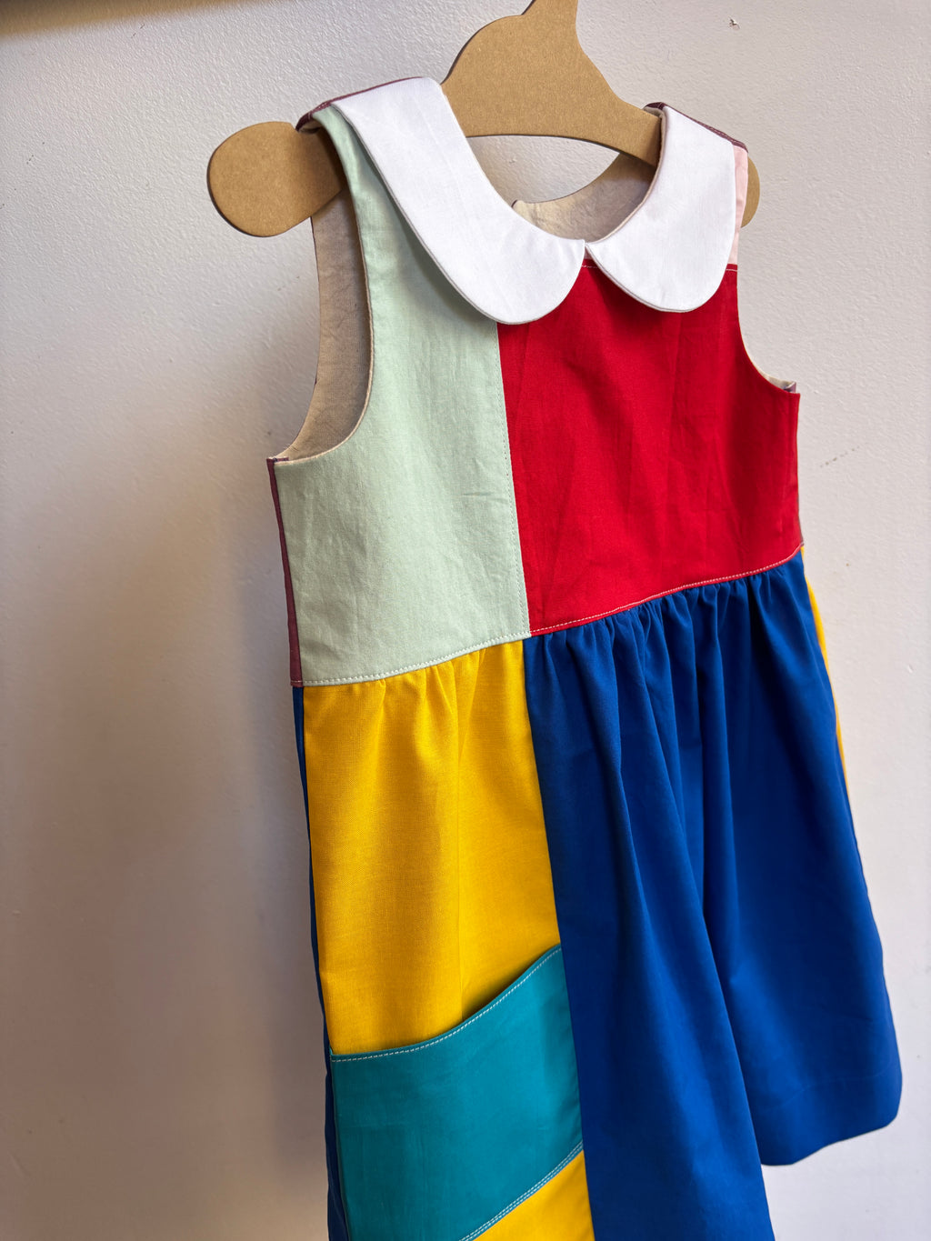 A sleeveless children's dress hanging on a wooden hanger against a plain wall. The dress has a Peter Pan collar and is made of large color-blocked fabric panels in white, red, yellow, blue, and teal.