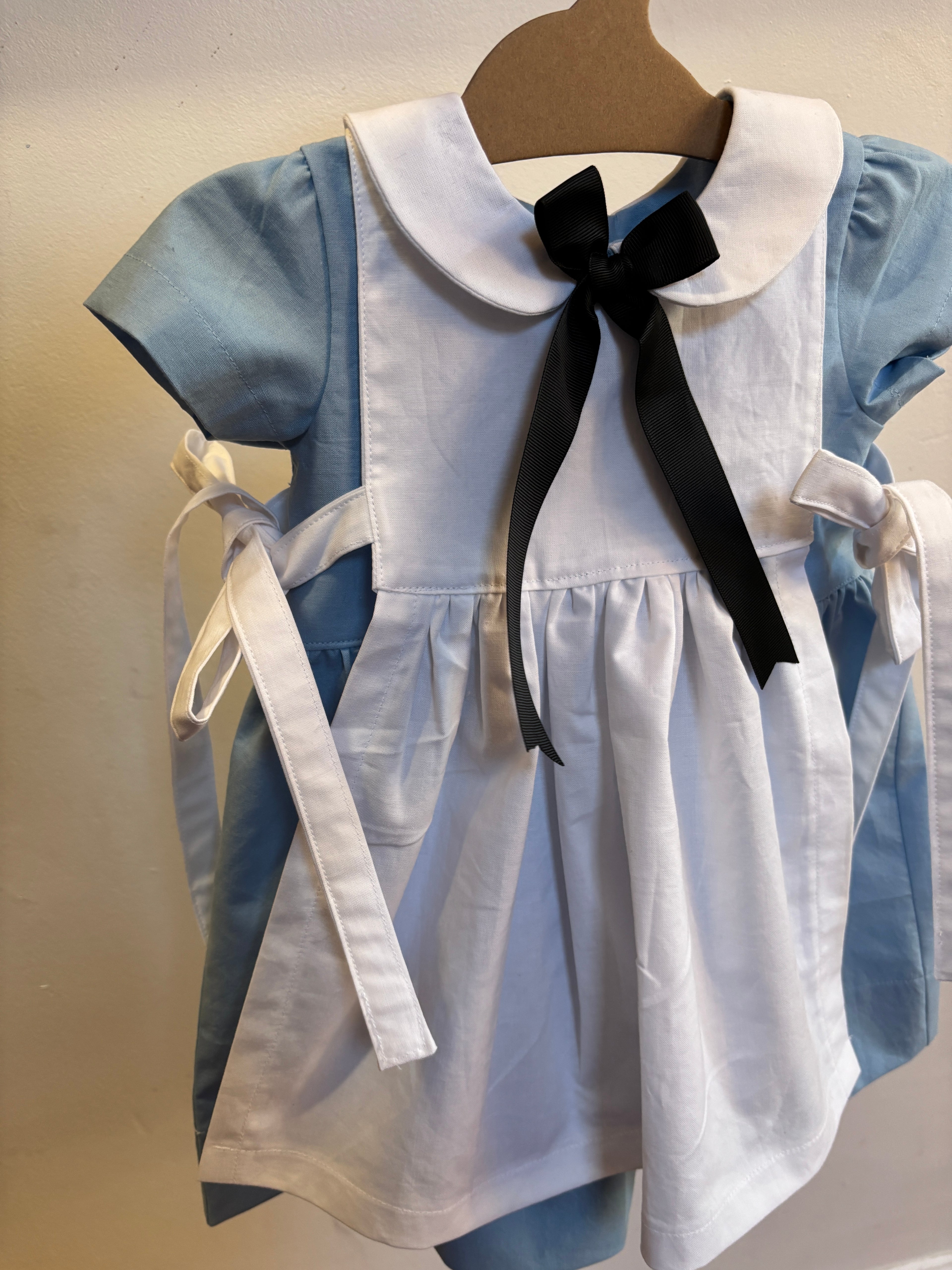 A toddler girl's dress featuring a white linen pinafore-style front with a large white Peter Pan collar and a black ribbon bow, layered over a light blue short-sleeve dress, hanging on a wooden hanger against a plain beige wall.
