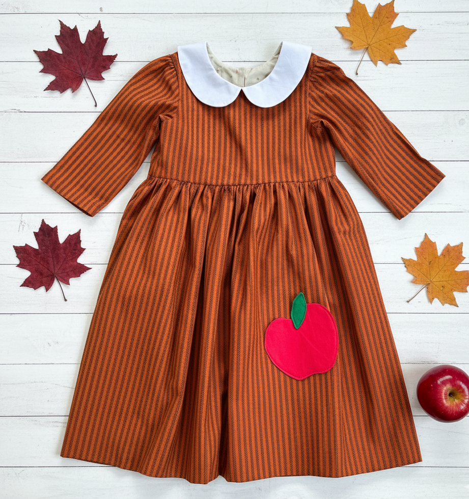 Brown striped dress with a red apple applique on a white wooden surface with autumn leaves.