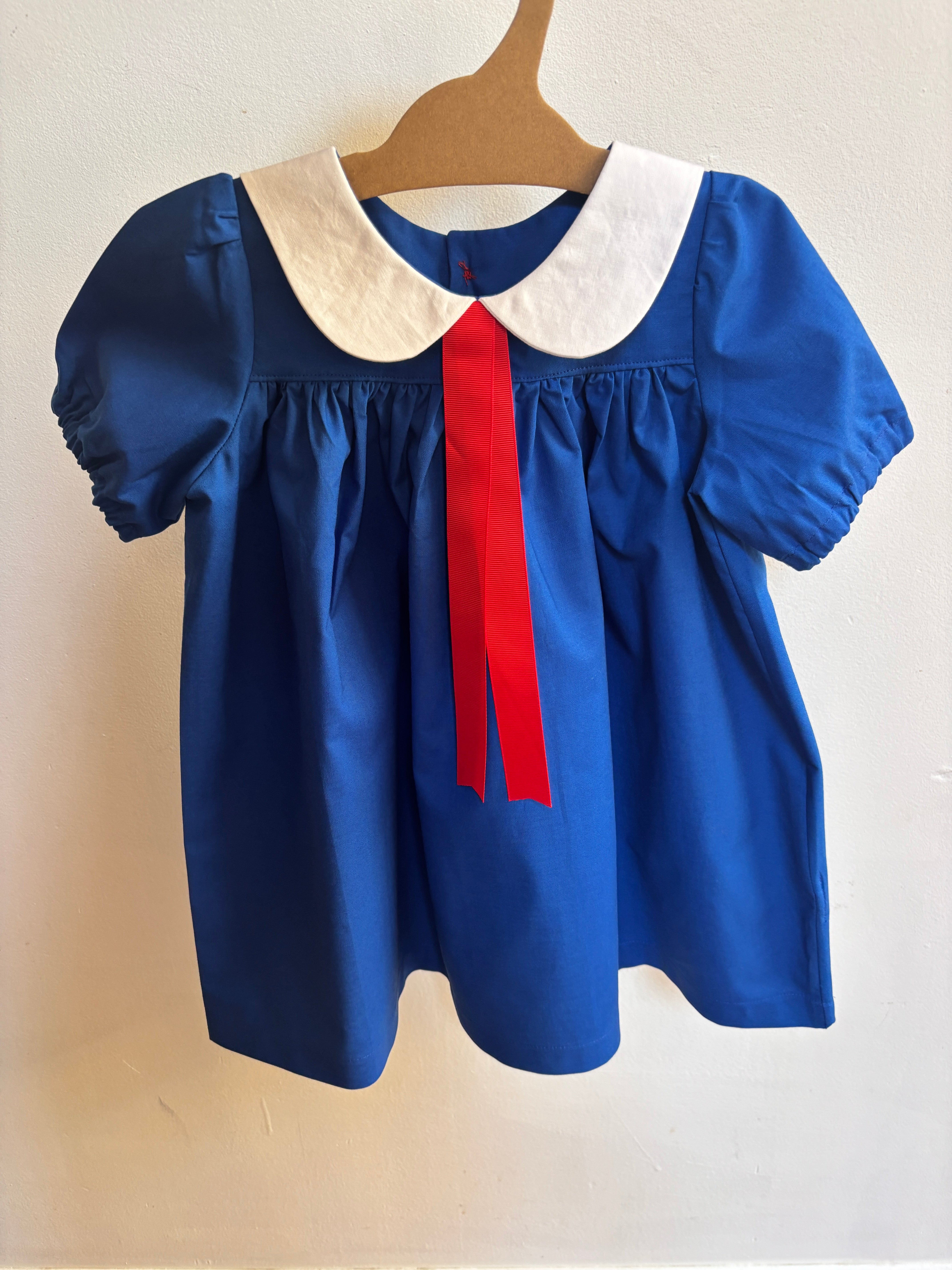 A blue short-sleeve dress with a white Peter Pan collar and a red ribbon tie, hanging on a wooden hanger against a plain off-white wall.