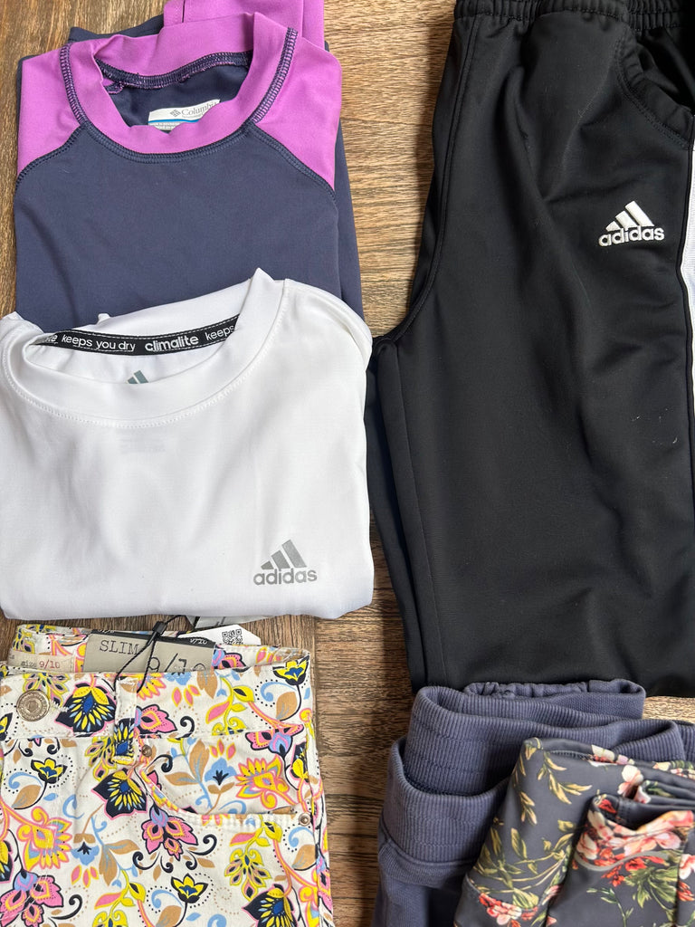 Adidas athletic wear including a purple and gray top, white shirt, black pants, and floral leggings on a wooden surface.