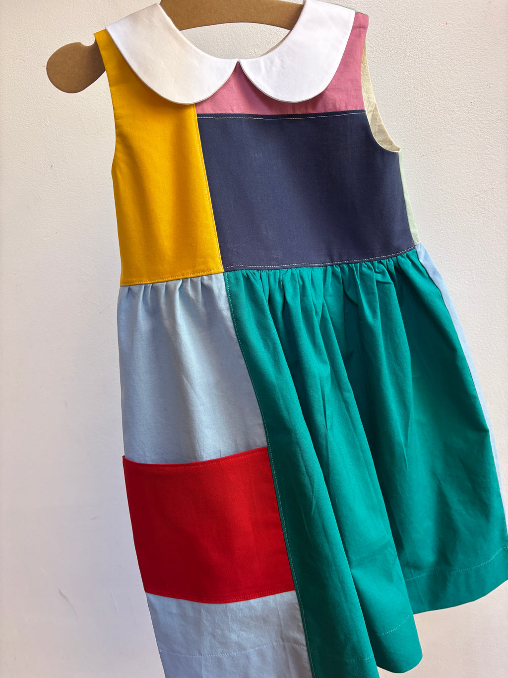 A sleeveless patchwork dress displayed on a wooden hanger against a plain off-white wall. The dress features a white Peter Pan collar, a yellow panel, a dark navy panel, a pink collar section, a light blue panel, a red band, and a large teal skirt.