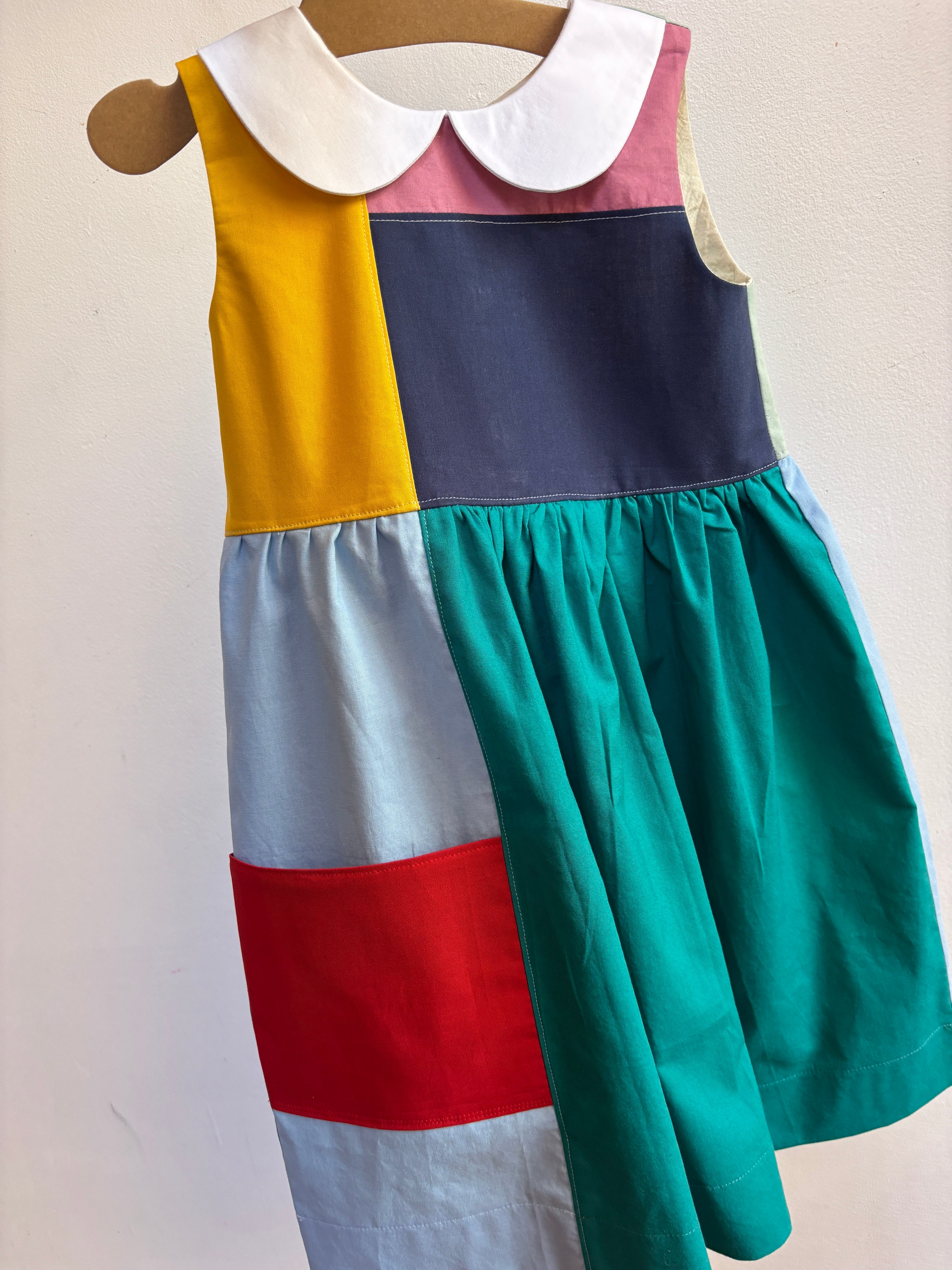 A sleeveless patchwork dress displayed on a wooden hanger against a plain off-white wall. The dress features a white Peter Pan collar, a yellow panel, a dark navy panel, a pink collar section, a light blue panel, a red band, and a large teal skirt.