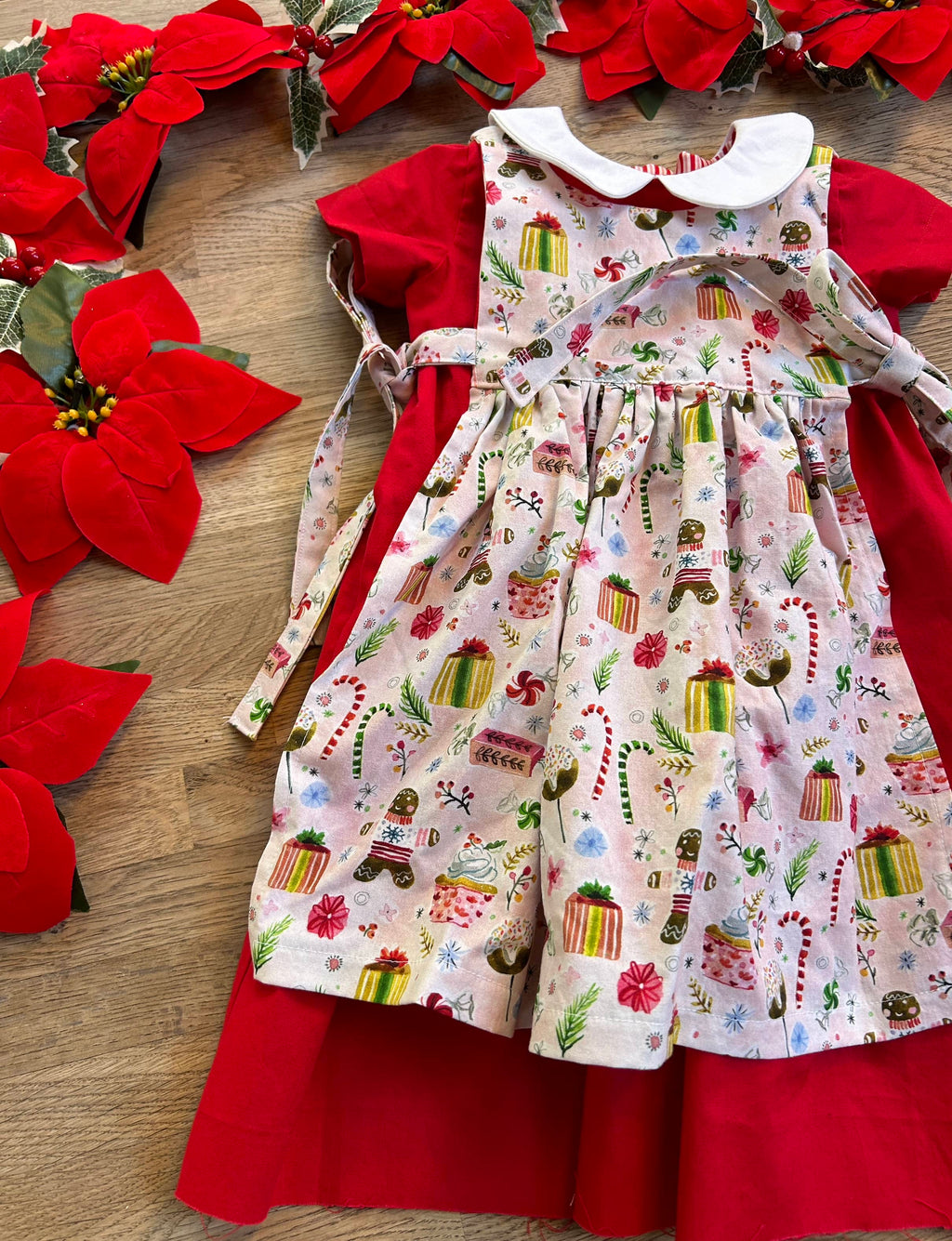 A red holiday dress with a floral and Christmas-themed pattern, laid out on a wooden surface with red poinsettias and green foliage around it.