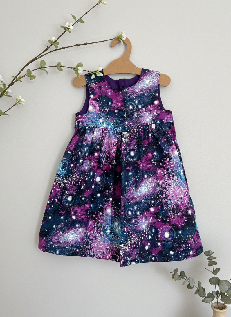 Children's dress with galaxy pattern on a wooden hanger against a light gray background.