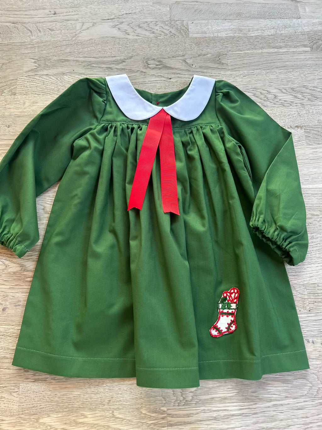 Green dress with a red bow and Christmas stocking patch on a wooden floor.