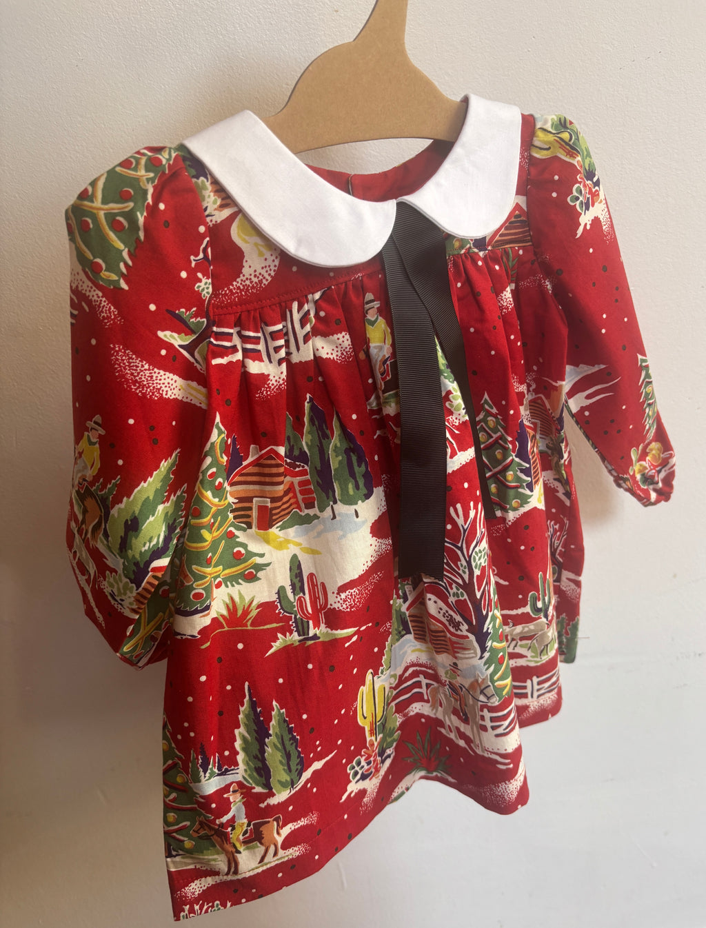 A baby girl's long-sleeve dress with a festive Christmas print on a red background, featuring a white Peter Pan collar and a black ribbon bow at the neckline, hanging on a wooden hanger against a plain wall.