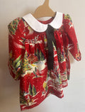 A baby girl's long-sleeve dress with a festive Christmas print on a red background, featuring a white Peter Pan collar and a black ribbon bow at the neckline, hanging on a wooden hanger against a plain wall.