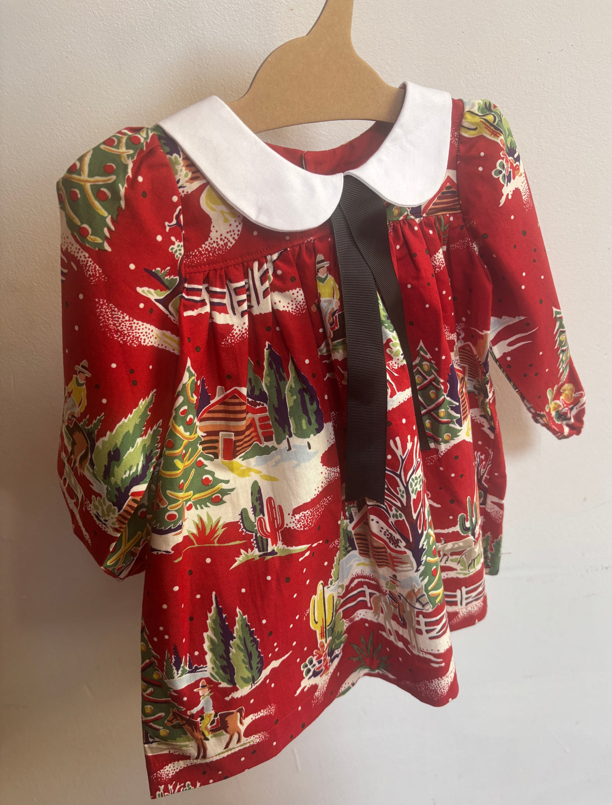 A baby girl's long-sleeve dress with a festive Christmas print on a red background, featuring a white Peter Pan collar and a black ribbon bow at the neckline, hanging on a wooden hanger against a plain wall.