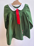 A green long-sleeve school uniform blouse with a white Peter Pan collar and a red neck ribbon, hanging on a wooden hanger against a plain white wall.