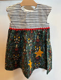 A sleeveless children's dress hanging on a wooden hanger against a plain off-white wall. The top portion has black and white horizontal stripes with a red scalloped trim, while the skirt features a dark green woodland-style print with yellow stars, red berries, and small illustrated motifs.