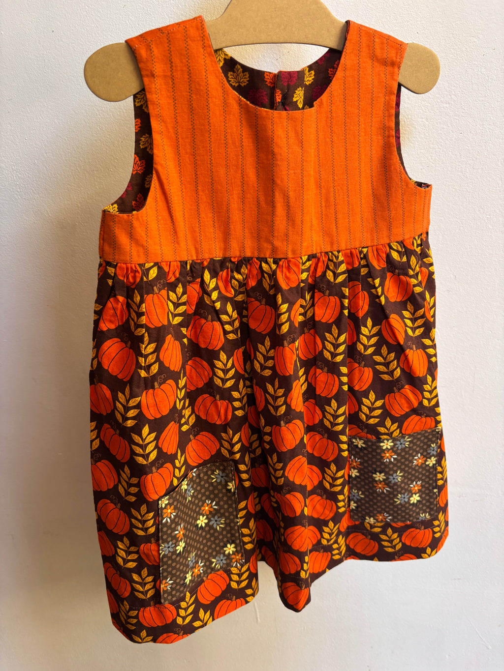 Little orange pumpkin dress hanging on a hanger with a white background. Dress has 2 brown pockets on the front skirt. 