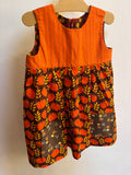 Little orange pumpkin dress hanging on a hanger with a white background. Dress has 2 brown pockets on the front skirt. 