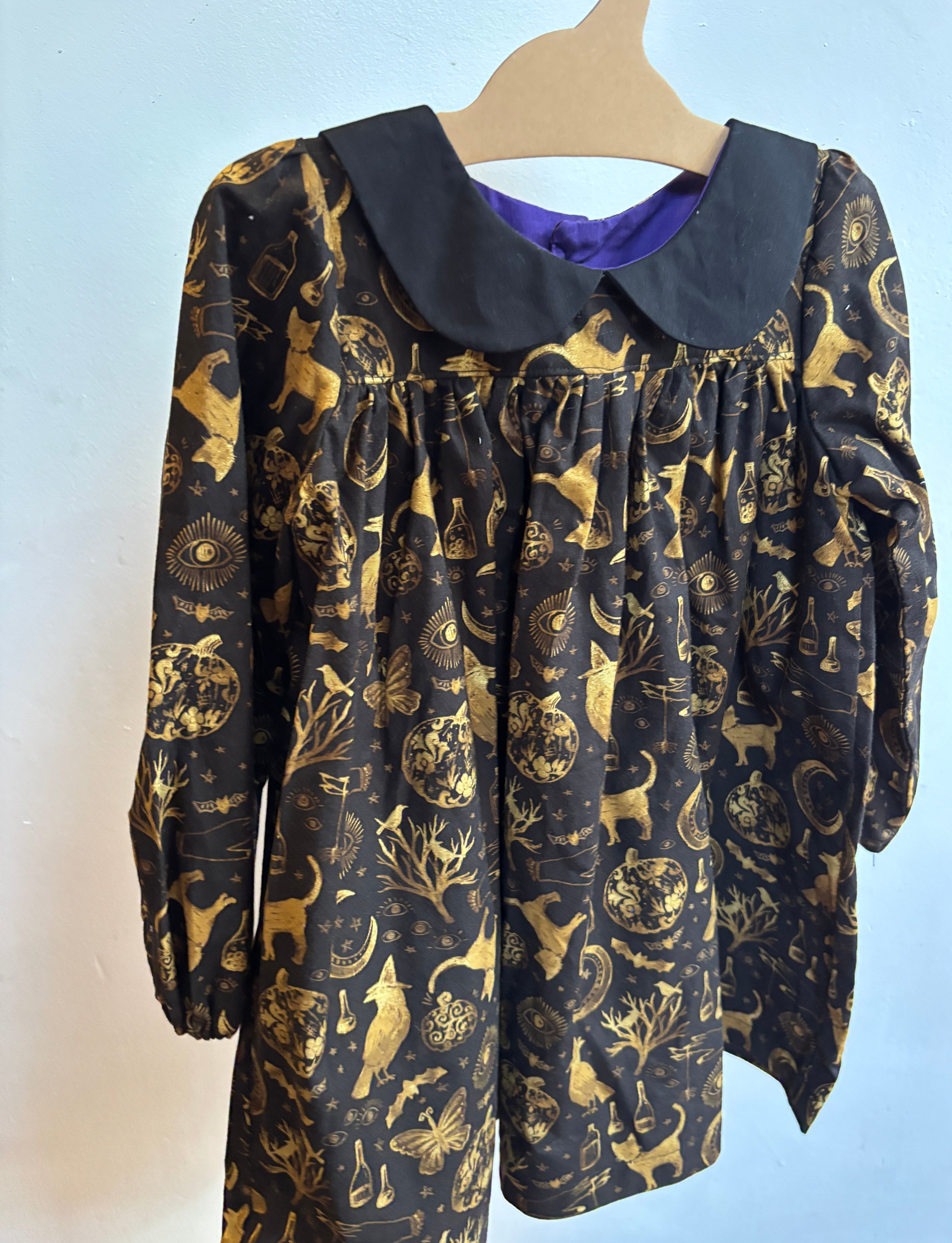 A long-sleeve baby dress with a black Peter Pan collar and a gold-toned occult-themed print featuring alchemical symbols, stars, and creatures, hanging on a wooden hanger against a light blue wall.