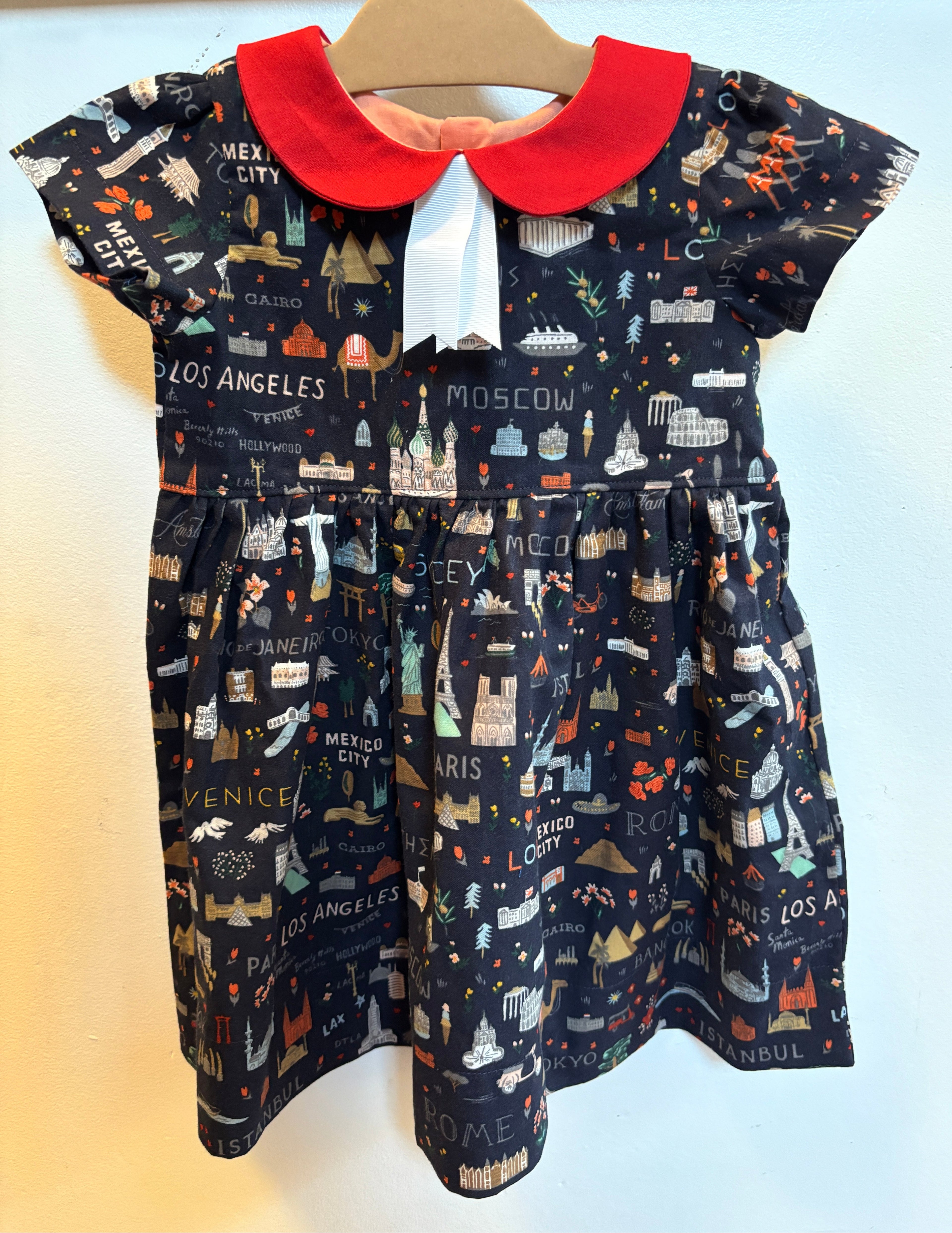 A toddler girl's short-sleeve dress hanging on a wooden hanger against a plain wall. The dress has a dark navy blue background printed with a whimsical cityscape pattern featuring illustrations of landmarks and city names like Los Angeles, Moscow, and Paris.