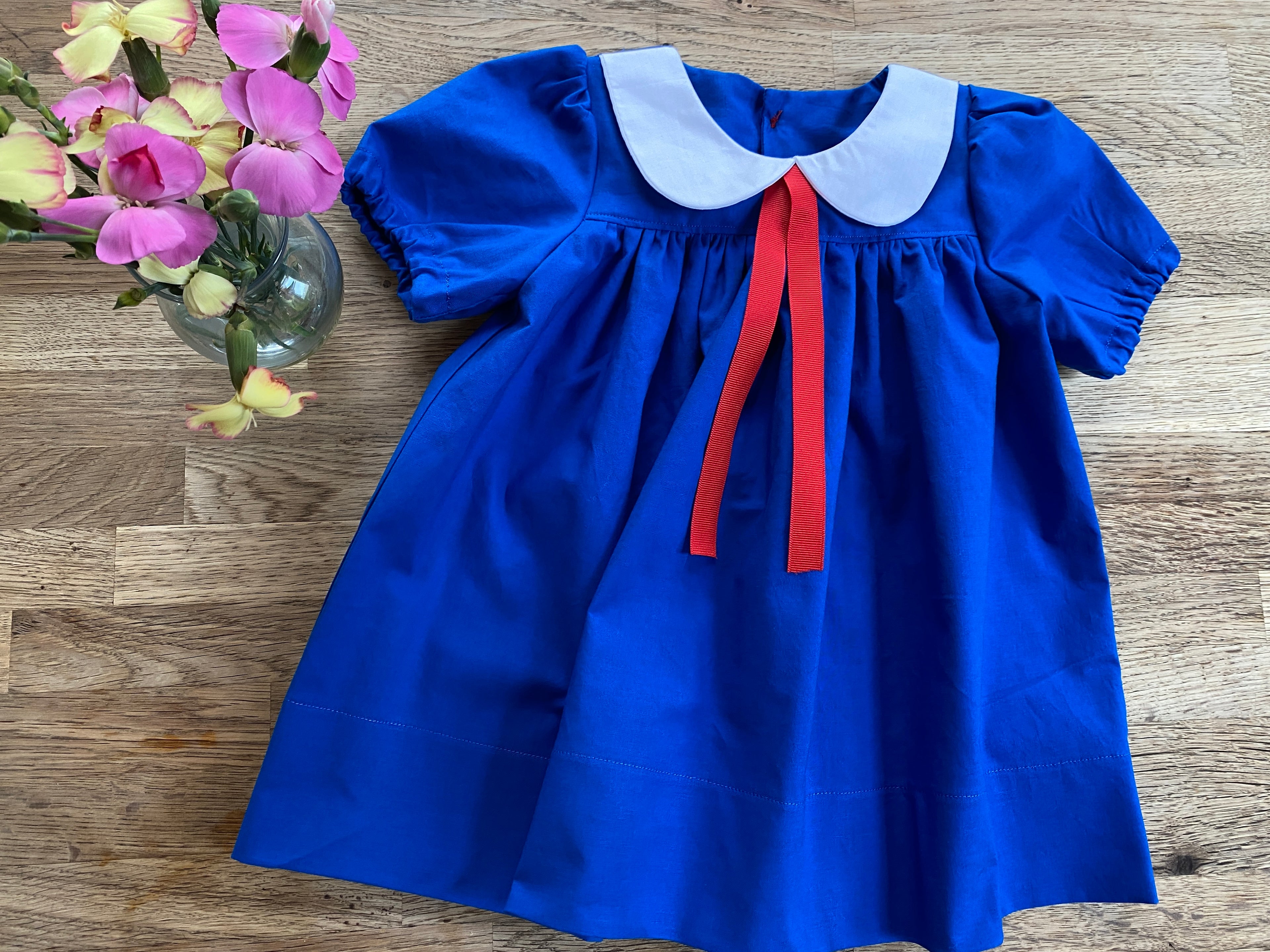 A vintage style royal blue dress with a white collar and a red ribbon bow at the neckline, displayed on a wooden surface with flowers in the background.