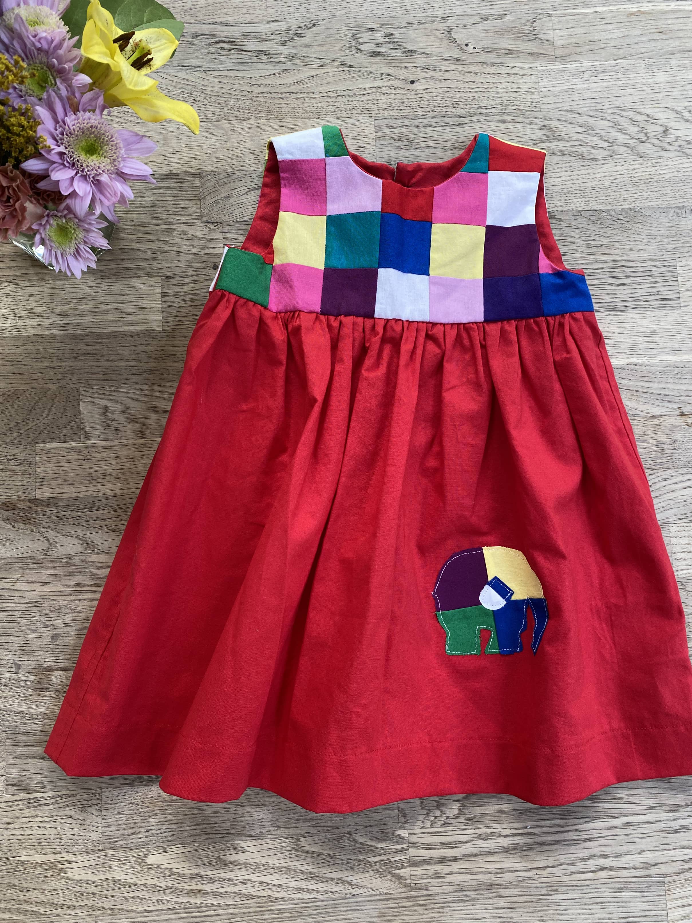 A colorful sleeveless patchwork dress for toddlers, featuring a red gathered skirt, a patchwork bodice with contrasting colors, and an appliquéd elephant on the front.